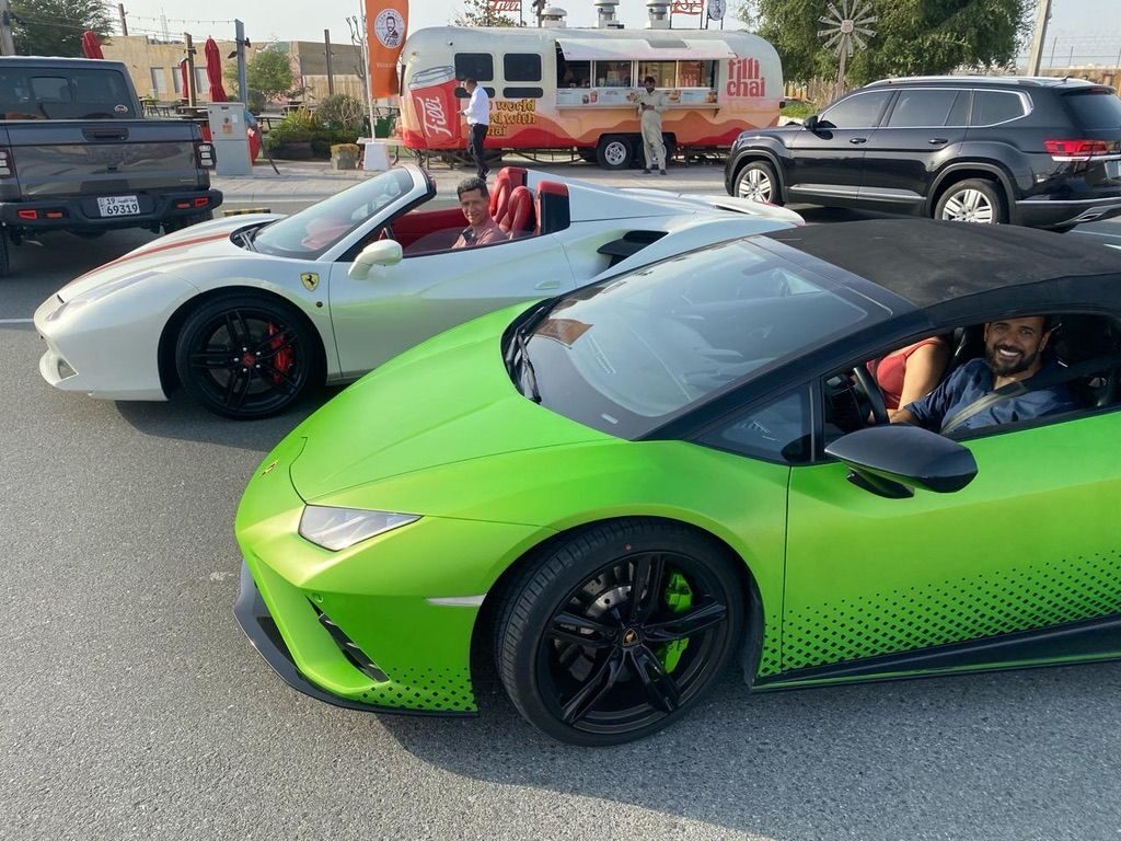 Ferrari branca e Lamborghini verde em movimento numa via com um food truck ao fundo